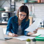 young woman working with computer while consulting some invoices and documents in the kitchen
